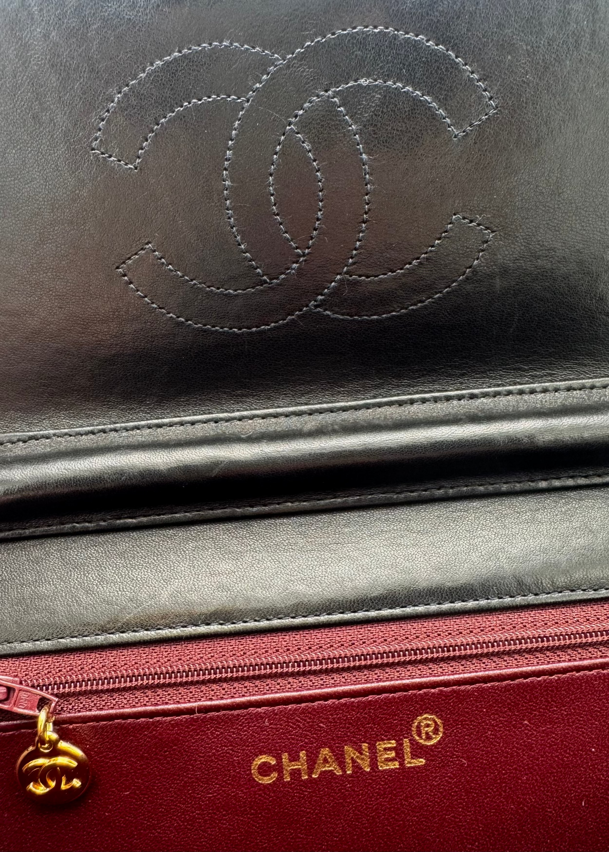 Close-up of a Chanel leather accessory showing the embossed interlocking CC logo in dark gray/black leather above a red zippered pocket with gold CHANEL lettering and a gold zipper pull.