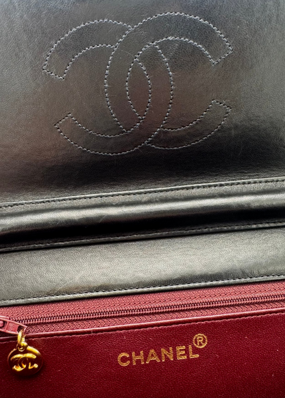 Close-up of a Chanel leather accessory showing the embossed interlocking CC logo in dark gray/black leather above a red zippered pocket with gold CHANEL lettering and a gold zipper pull.