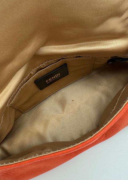Close-up of the interior of a Fendi bag, showing a gold satin lining with a black Fendi logo patch and an orange leather exterior edge visible at the bottom.