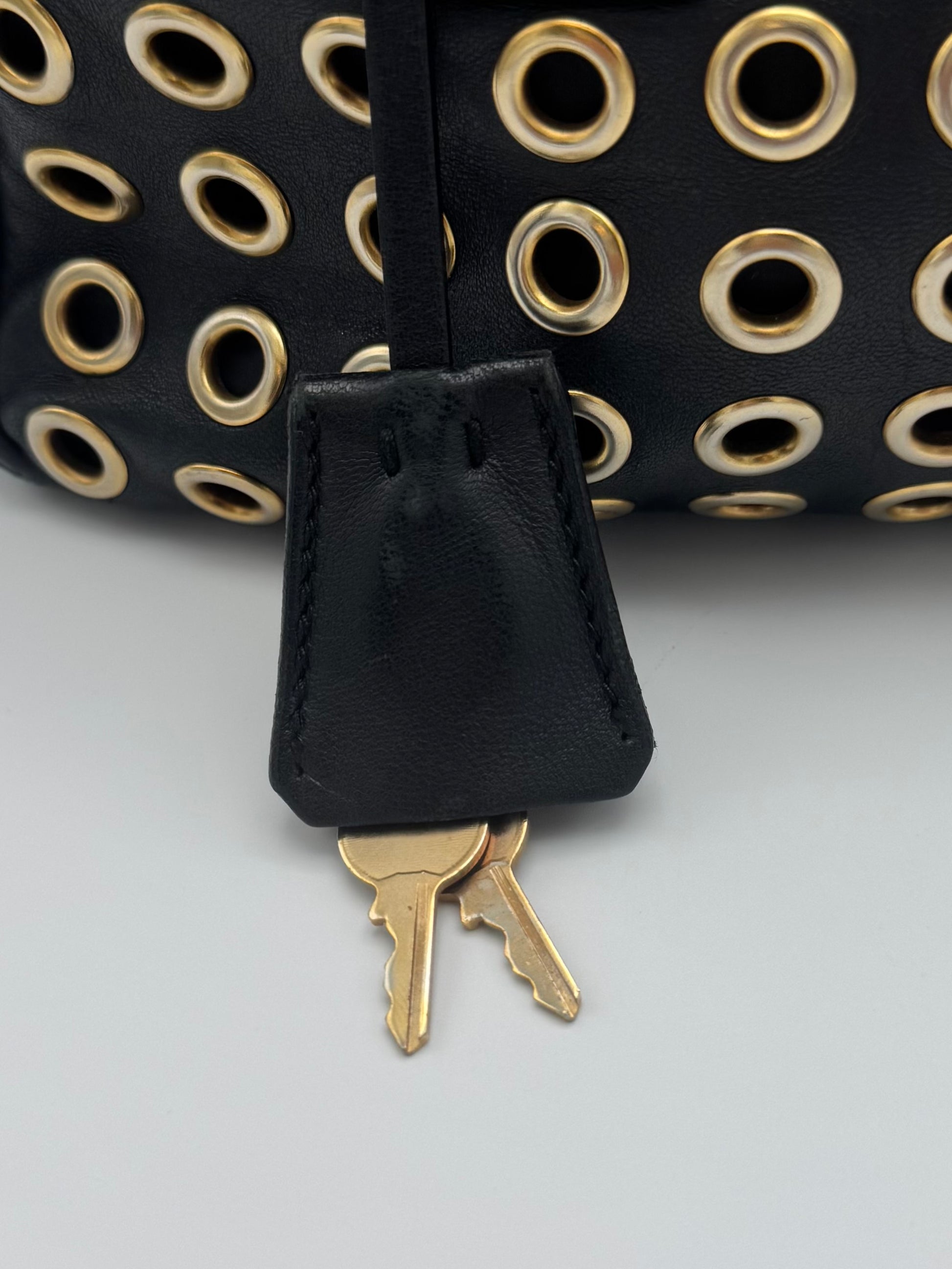Close-up of a black leather key fob accessory with gold-tone metal grommets and two keys attached, shown against a light gray background.