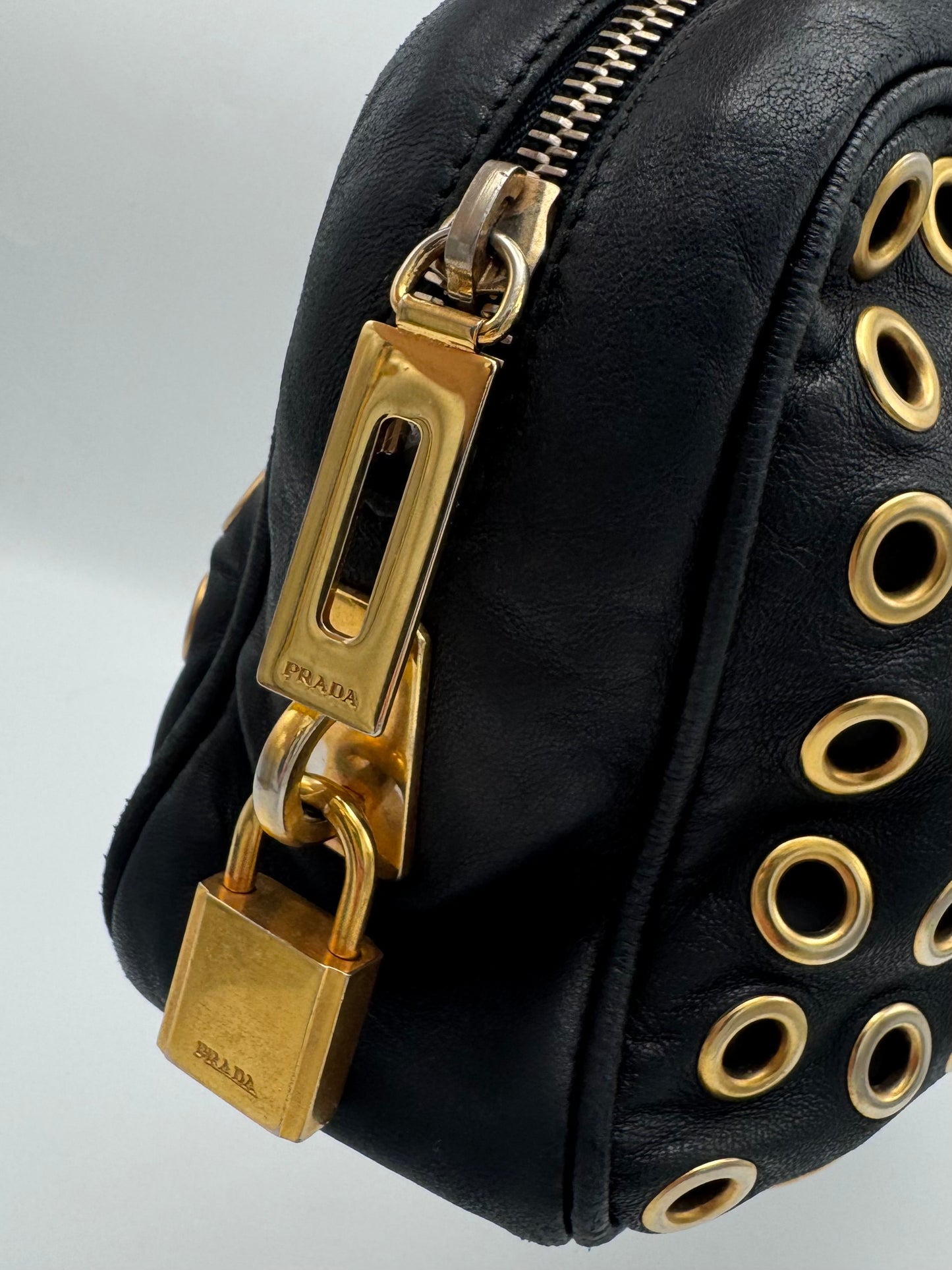 Close-up of a black leather Prada bag featuring gold-tone hardware, including a padlock charm, zipper pull, and circular grommet details on the side.