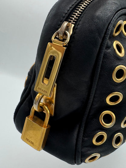 Close-up of a black leather Prada bag featuring gold-tone hardware, including a padlock charm, zipper pull, and circular grommet details on the side.