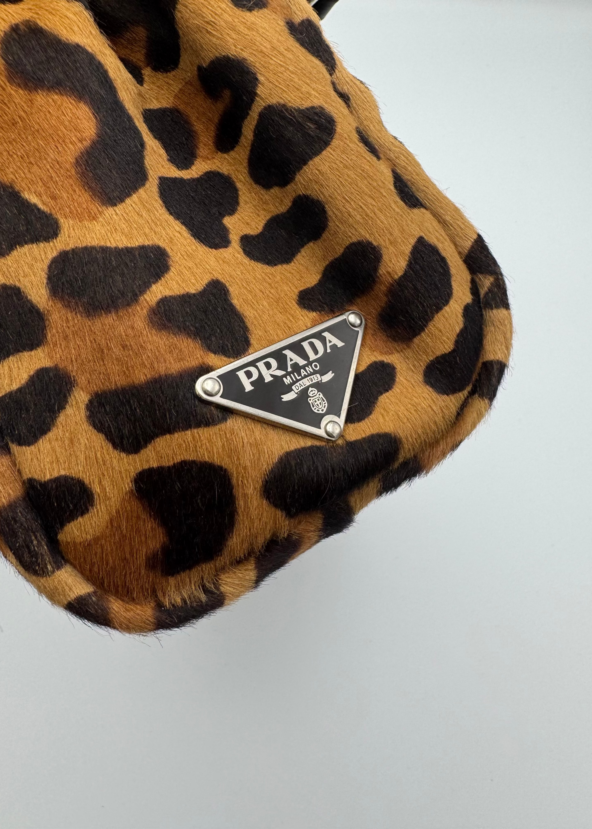 Close-up of a Prada handbag in leopard print fur with a silver-tone triangular Prada logo plaque on a light gray background.