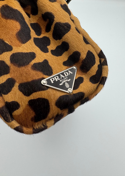 Close-up of a Prada handbag in leopard print fur with a silver-tone triangular Prada logo plaque on a light gray background.