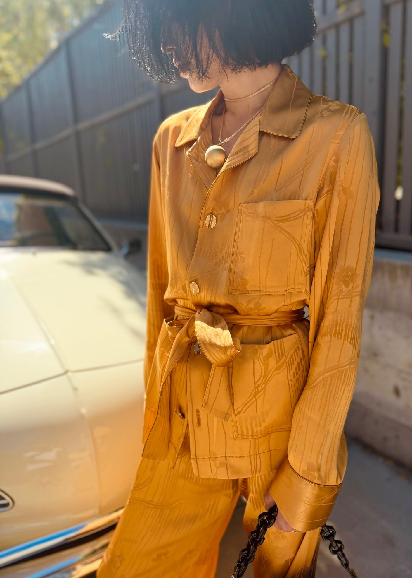 A person wearing a forte_forte mustard yellow matching shirt and pants set with a subtle vertical textured stripe pattern, standing outdoors next to a vintage cream-colored car.